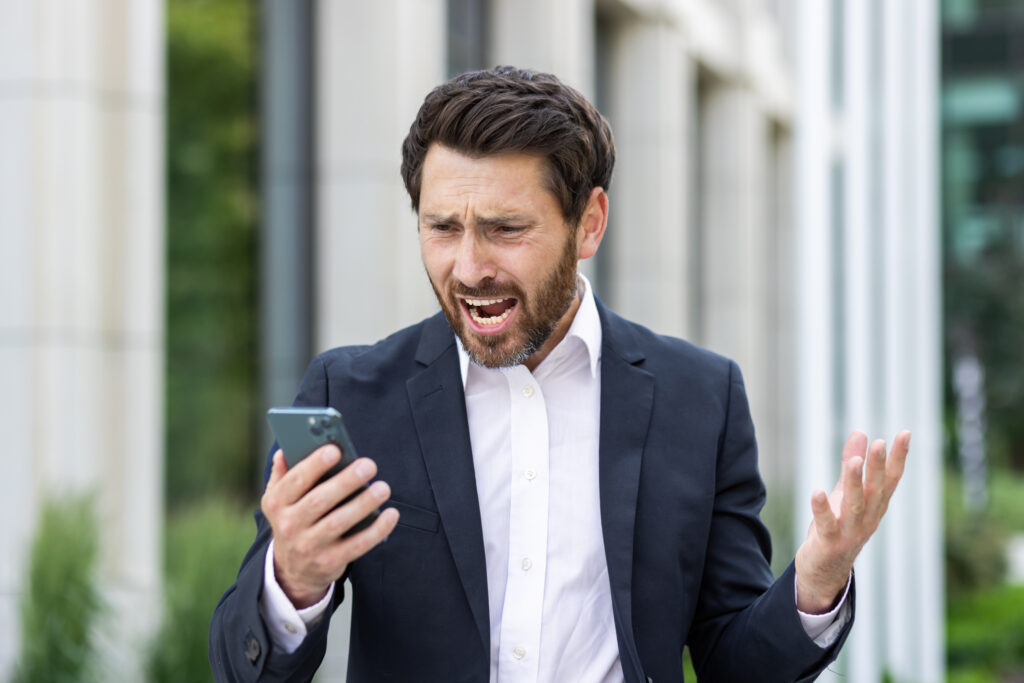 Üzletember sötét öltönyben, aki dühösen nézi a telefonját. A kép az informatikai problémák okozta stresszt és frusztrációt illusztrálja.