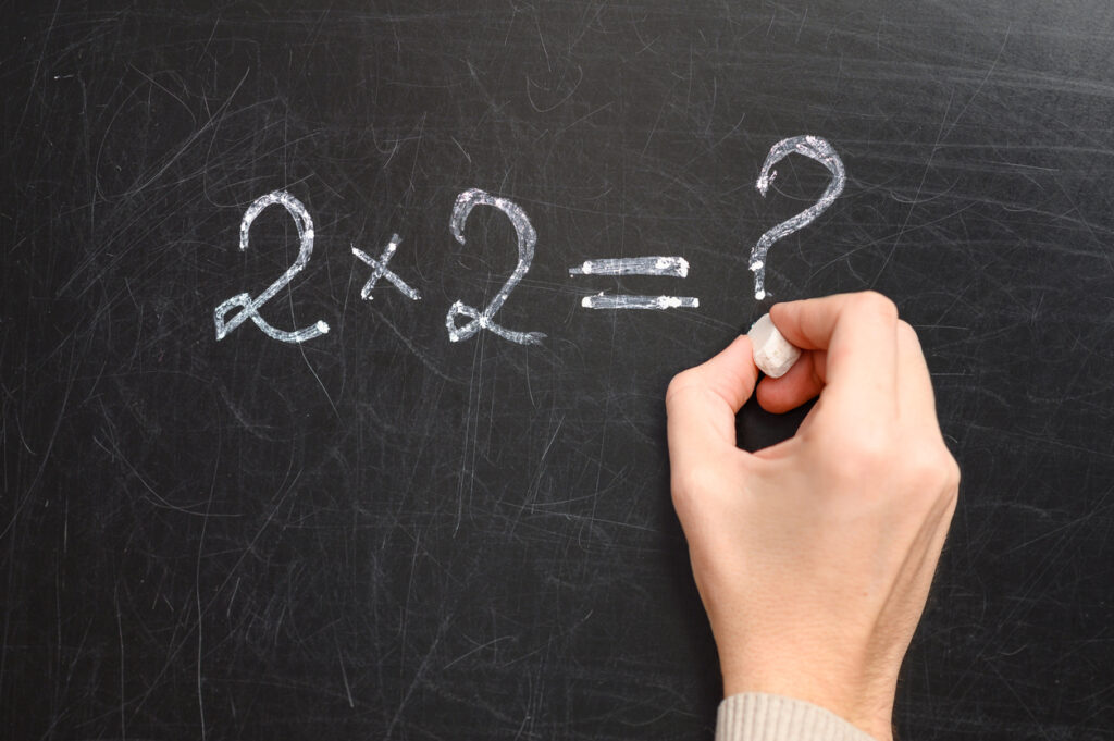Egy kéz krétával egy iskolai táblára írja a 2x2=? matematikai kifejezést, oktatás és kérdésfelvetés szimbolikája.