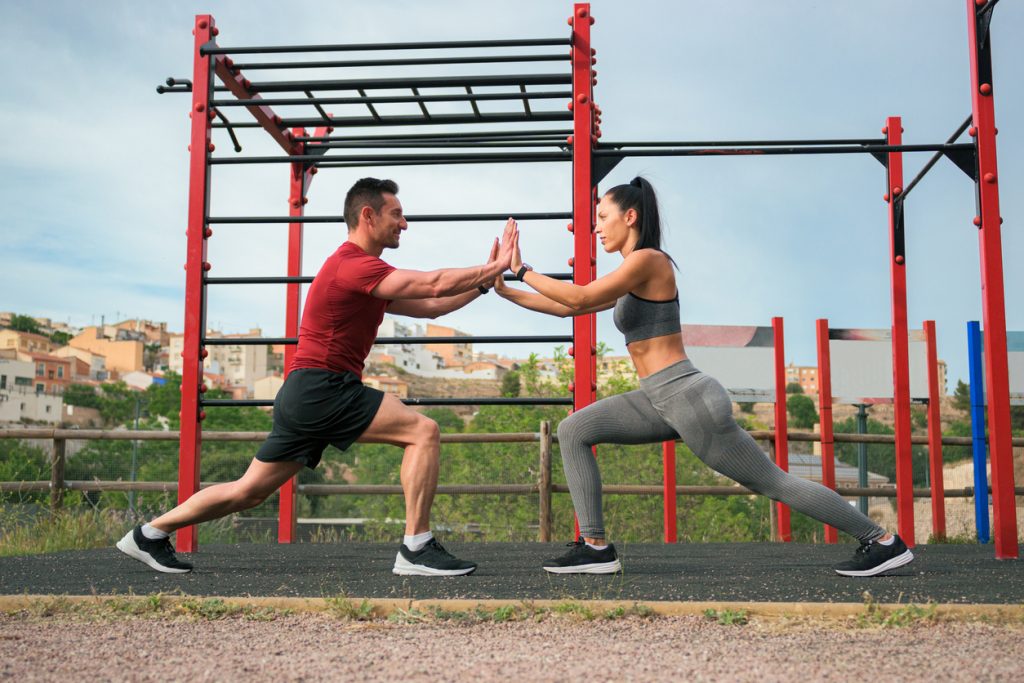 Két ember közösen edz egy szabadtéri fitneszparkban, testmozgás és együttműködés szimbóluma, városi háttérrel.