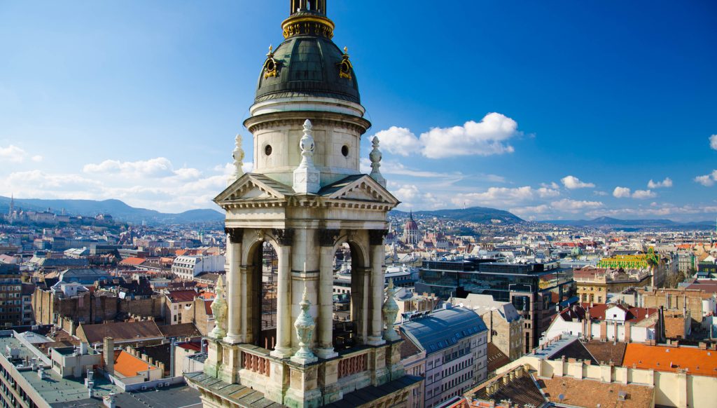 Budapesti panoráma a Szent István-bazilika tornyával az előtérben, kék égbolttal és városi látképpel.